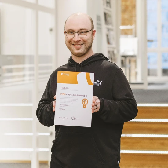 T wie Tim und TYPO3 Foto unseres Kollegen Tim. Er hält stolz sein TYPO3 Zertifikat und lächelt in die Kamera.