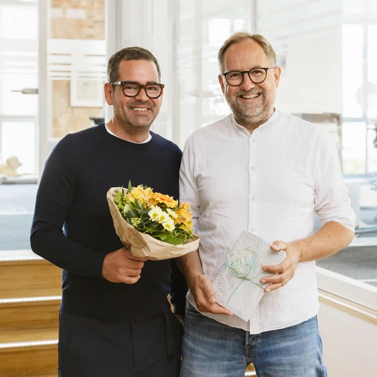 15 humorvolle Jahre mit Björn Foto von Chef Felix mit Standortleiter, Projektleiter und Prokurist Björn mit Blumen und einem Geschenk in den Händen