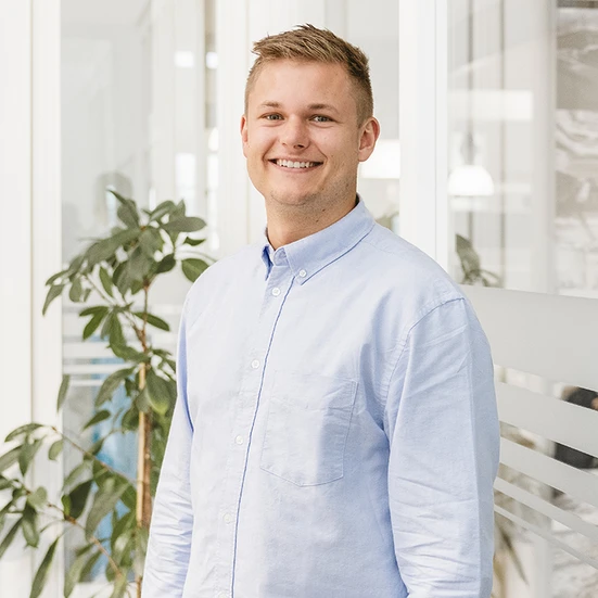 Tom Cordes Porträtfoto von Tom er trägt ein hellblaues Hemd und lächelt freundlich in die Kamera. Im Hintergrund Glaswände der Büroräume und eine Pflanze.